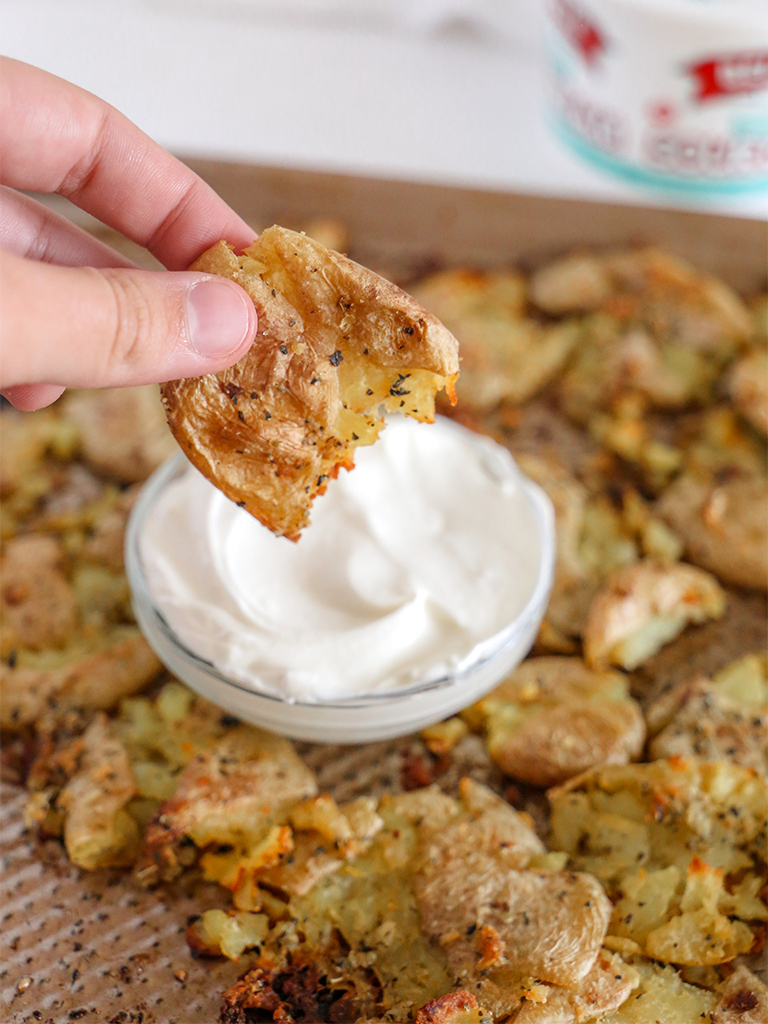 A hand dipping a piece of garlic butter smashed potato into a bowl of sour cream, with more smashed potatoes and sour cream container in the background.