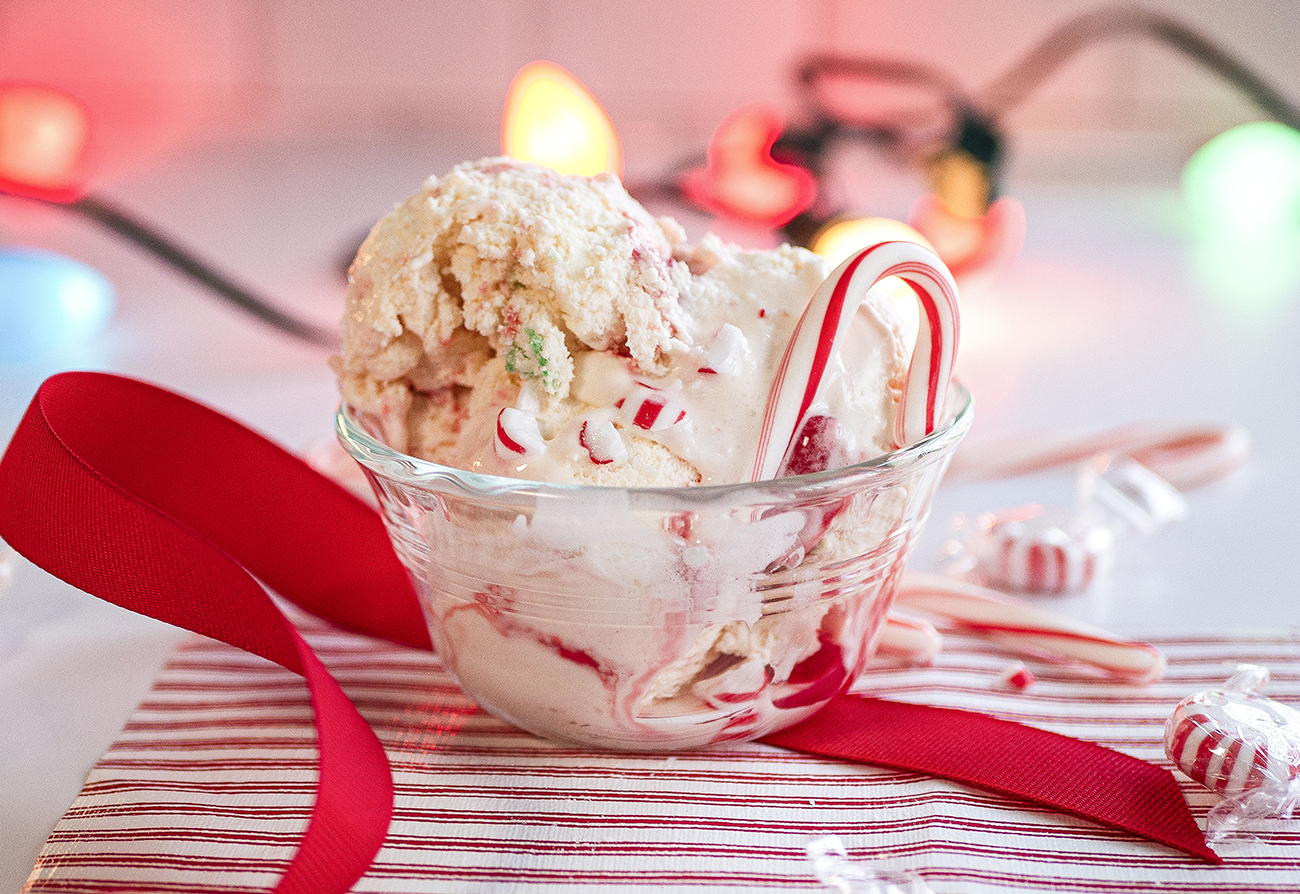 A festive bowl of peppermint ice cream with a candy cane sits on a striped cloth, surrounded by lights and a red ribbon.