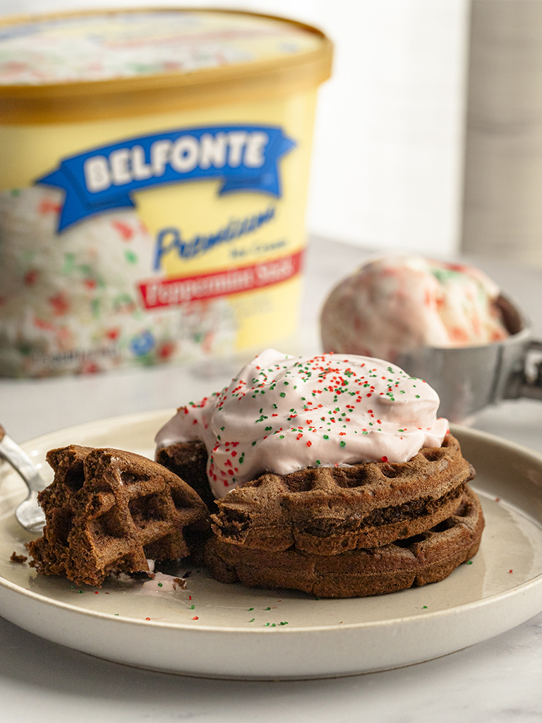 Chocolate waffles stacked on a plate, topped with Belfonte Peppermint Stick whipped ice cream and festive red and green sprinkles.