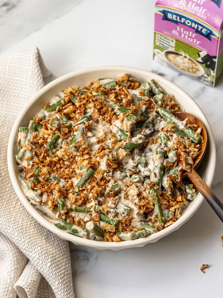 A creamy green bean casserole topped with crispy fried onions, served in a white dish, with a carton of Belfonte Dairy Half and Half in the background.