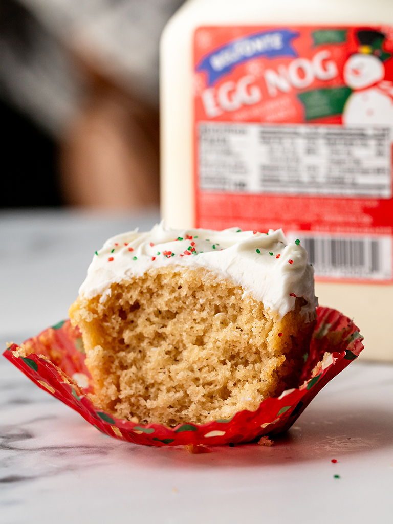 A festive eggnog cupcake with creamy white frosting, sprinkles, and a bite taken out, showcasing its moist interior, with a container of Belfonte Eggnog in the background.