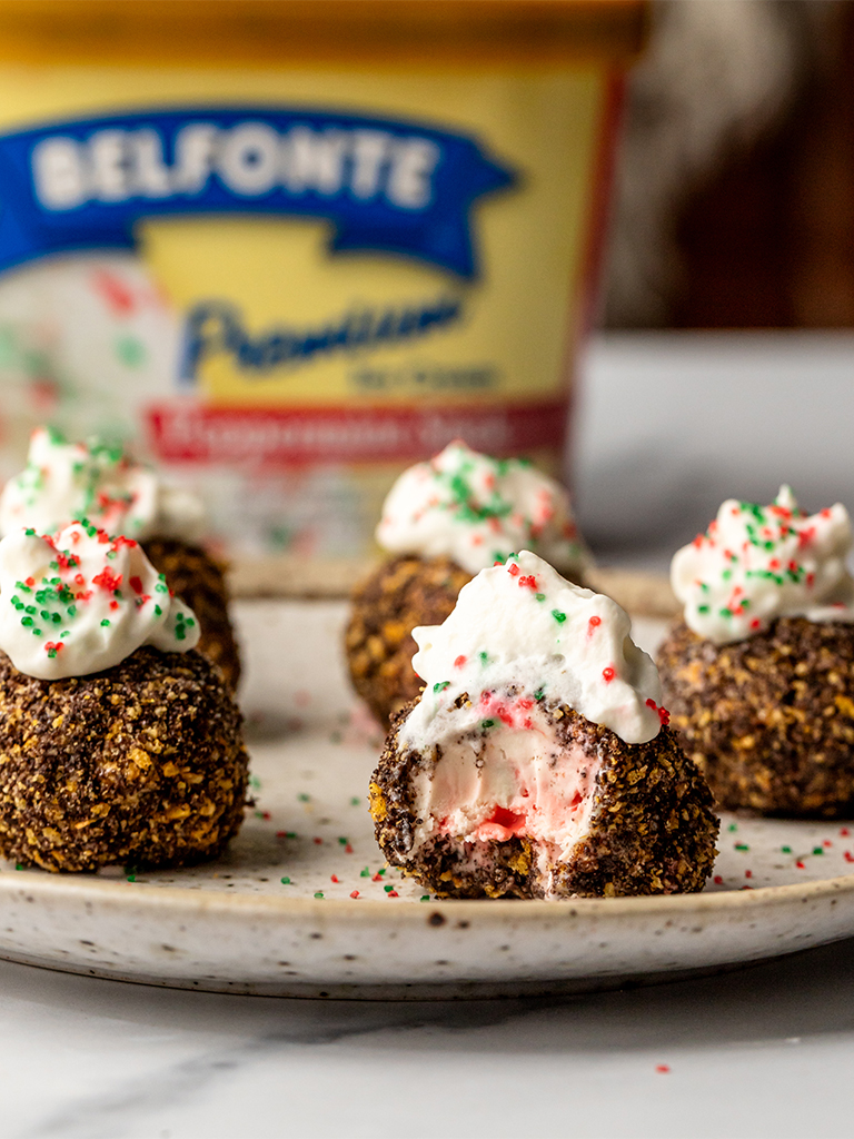 Close-up of fried ice cream bites on a plate, one with a bite taken out revealing pink peppermint ice cream inside, topped with whipped cream and holiday-colored sprinkles, with Belfonte Peppermint Ice Cream in the background.