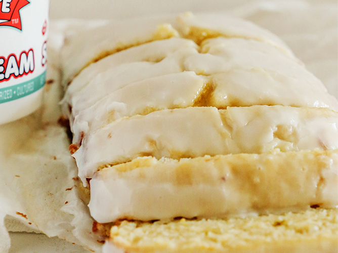 Sliced lemon loaf cake covered in a thick lemon glaze sits beside a container of sour cream, highlighting a zesty homemade treat.