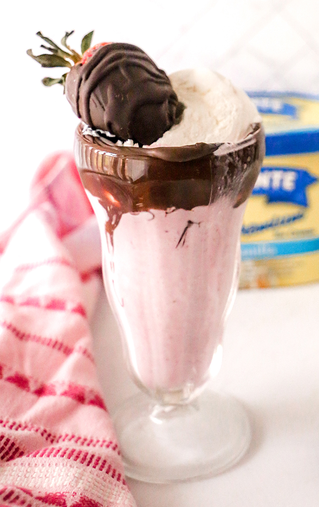 A chocolate-covered strawberry milkshake in a glass with a chocolate-drizzled rim, garnished with whipped cream and a chocolate-dipped strawberry, with a striped towel beside it.