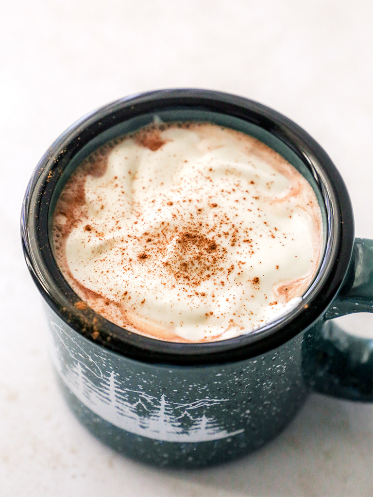 A warm mug of hot cocoa topped with whipped cream and sprinkled with cinnamon, served in a dark green mug featuring a winter forest design.