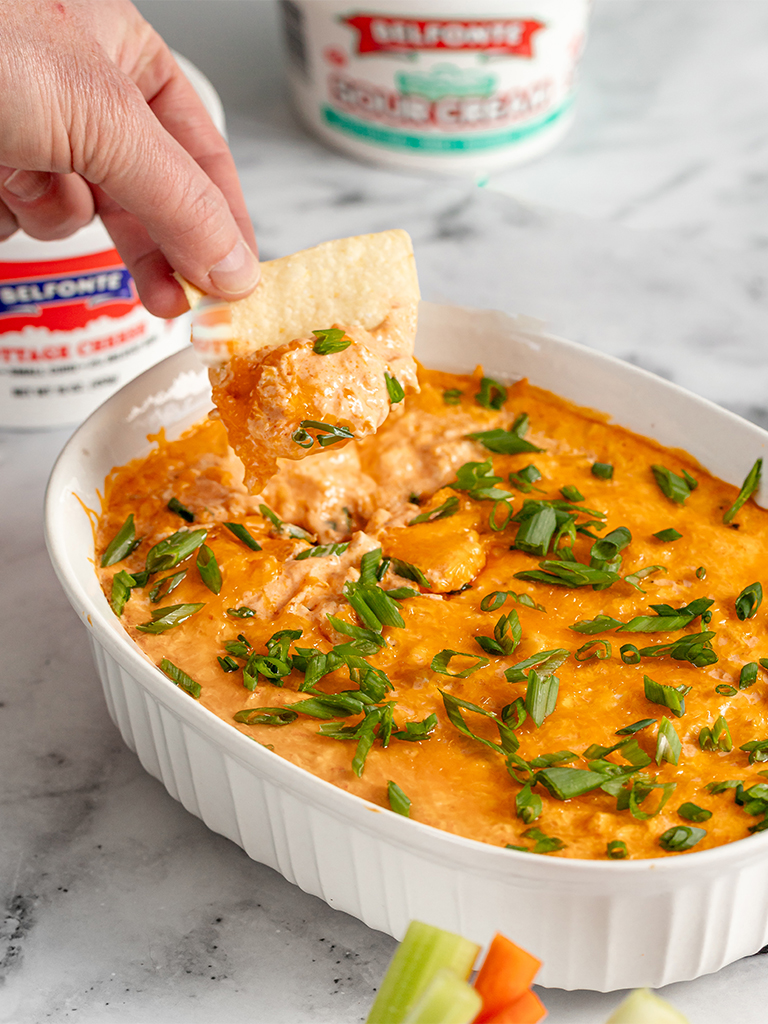 A creamy Buffalo Chicken Dip in a white casserole dish, topped with melted cheese and chopped green onions. A hand dips a tortilla chip into the cheesy mixture. In the background, containers of Belfonte Cottage Cheese and Sour Cream are visible.