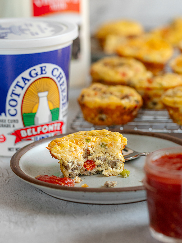 A partially eaten savory egg muffin with sausage, peppers, and cheese sits on a plate beside a jar of salsa, with a container of Belfonte Cottage Cheese in the background.