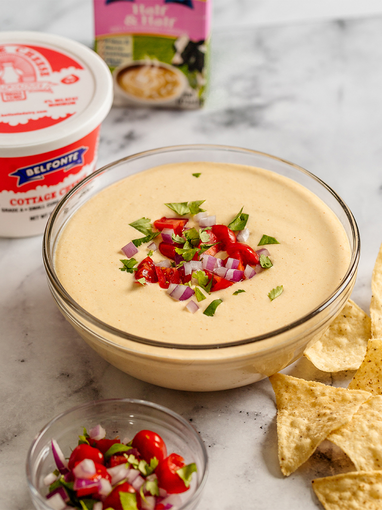 A glass bowl filled with creamy queso dip topped with diced red tomatoes, chopped red onion, and fresh cilantro. In the background, a container of Belfonte Cottage Cheese is slightly blurred. Tortilla chips are placed around the bowl, ready for dipping.