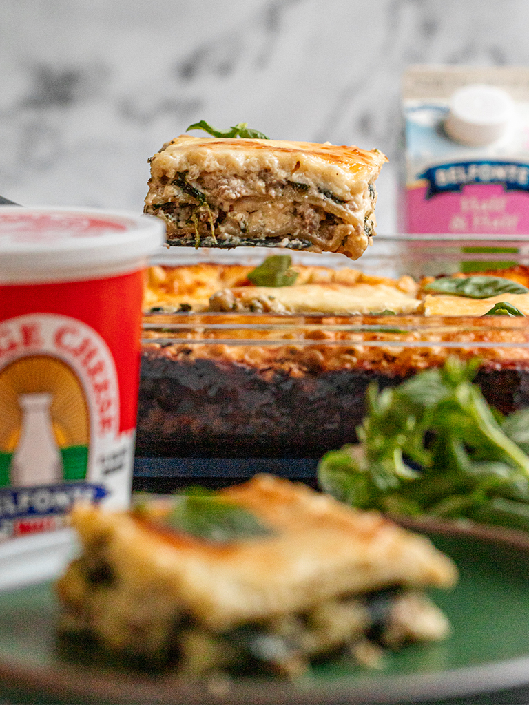 A slice of lasagna layered with spinach, Italian sausage, and cheese is lifted from a baking dish, with Belfonte Cottage Cheese and fresh herbs in the background.