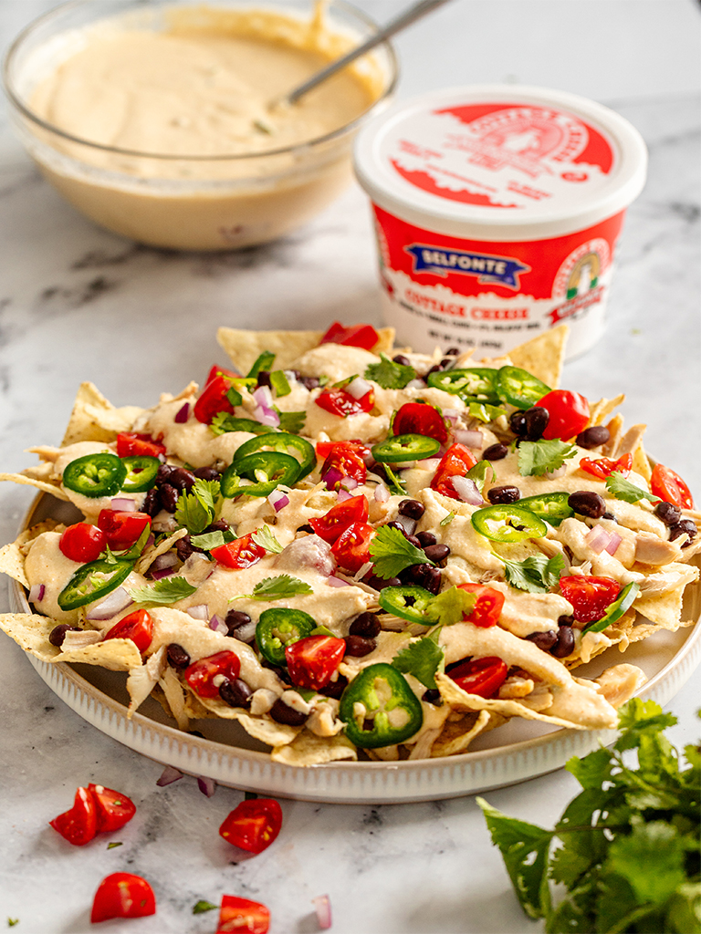 Cottage cheese queso nachos topped with black beans, diced tomatoes, jalapeño slices, red onion, and fresh cilantro on a white plate, with a container of Belfonte Cottage Cheese and a bowl of queso in the background.