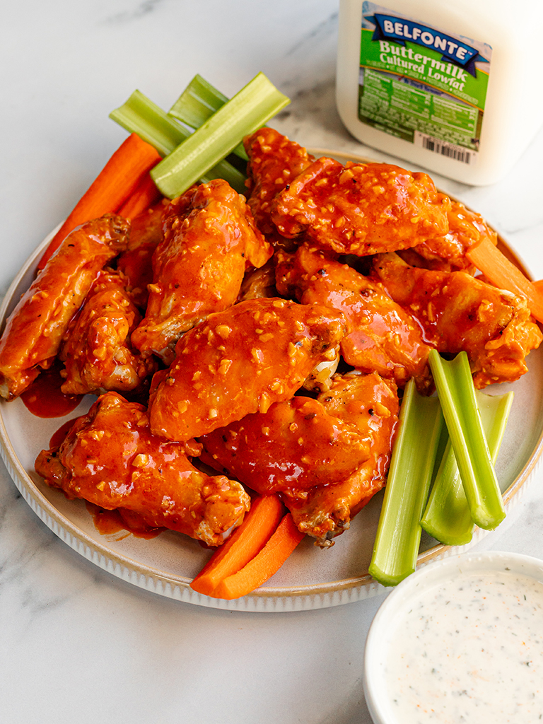 A close-up of buffalo chicken wings drenched in a thick, garlicky hot sauce, arranged on a plate with fresh celery and carrot sticks. A container of Belfonte Dairy Buttermilk is seen in the background.