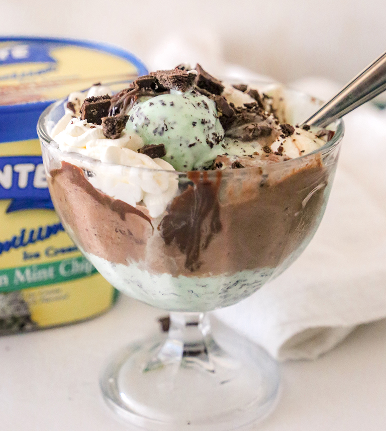 A glass dessert dish filled with a mint chocolate ice cream sundae topped with whipped cream, chocolate sauce, and crushed chocolate cookies. A container of Belfonte Premium Green Mint Chip ice cream is visible in the background.