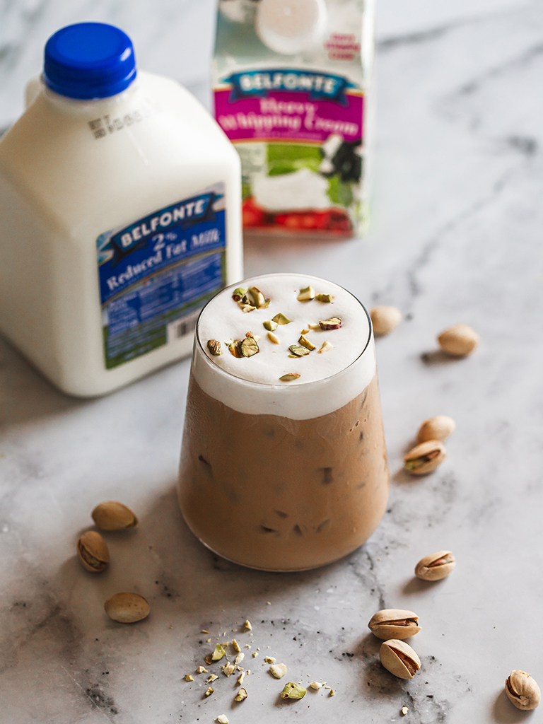 A glass of iced coffee topped with thick pistachio cold foam and garnished with crushed pistachios, surrounded by scattered nuts on a marble surface. In the background, containers of Belfonte 2% Reduced Fat Milk and Heavy Whipping Cream are visible.